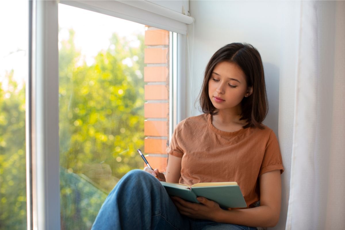 Young woman journaling by the window, reflecting on her thoughts and emotions
