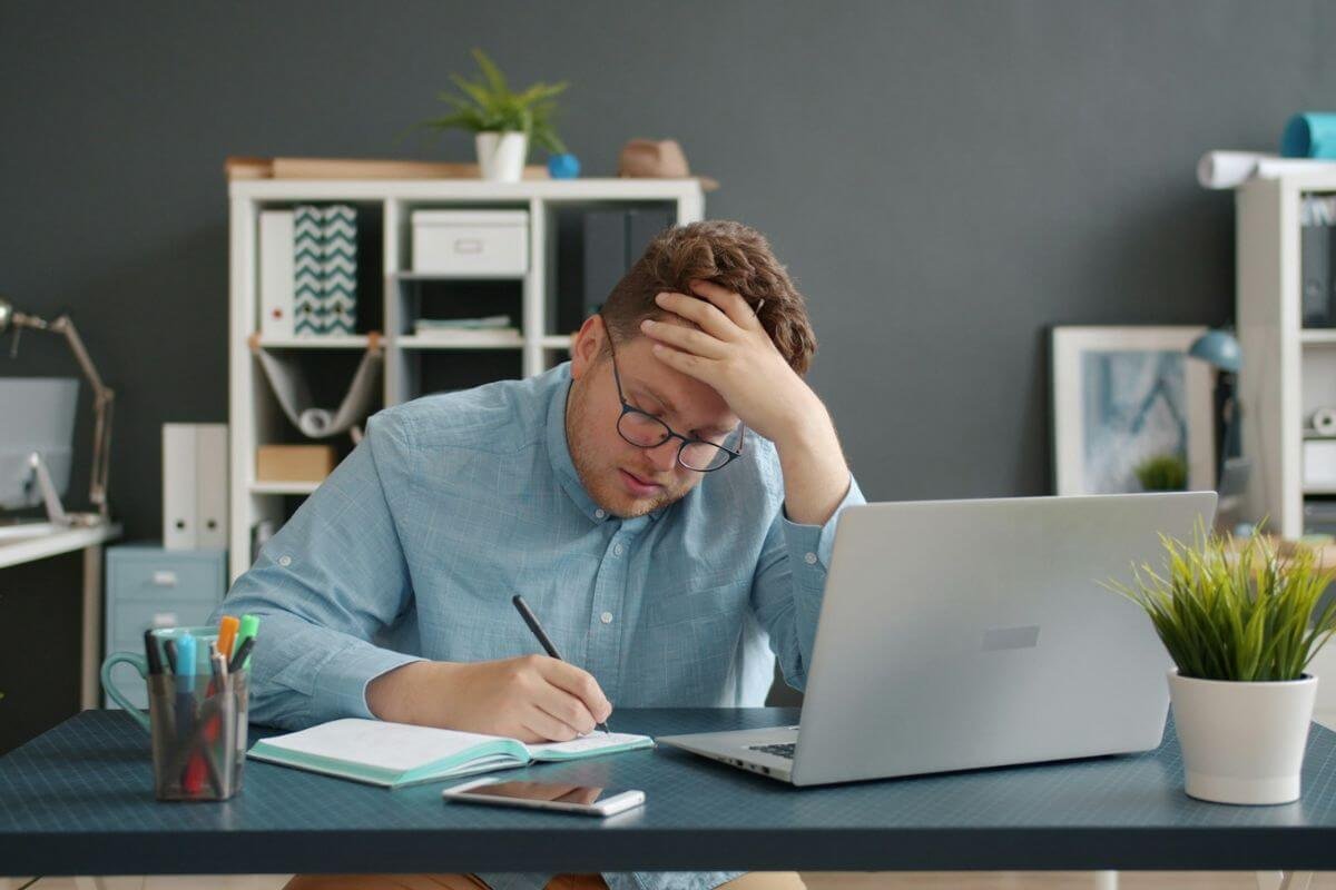 Stressed man holding his head while working on a laptop, symbolizing financial overwhelm and low money vibration.
