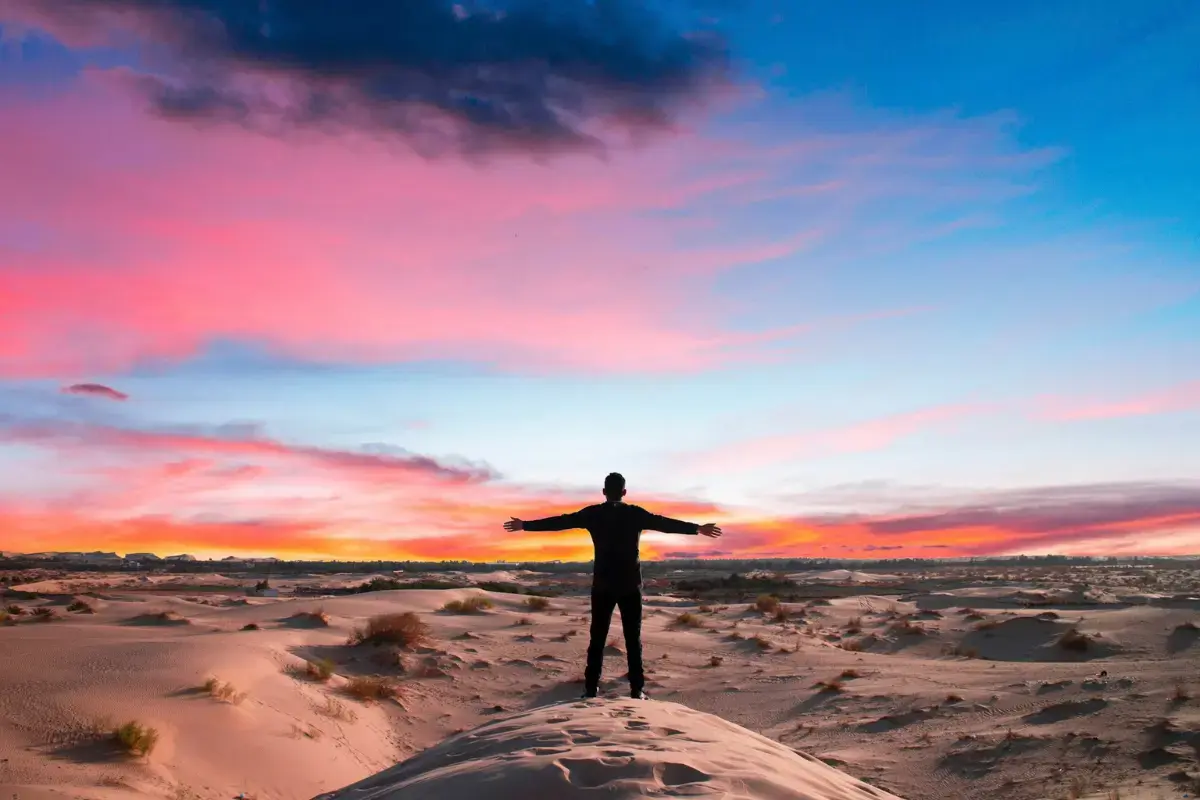 A man standing on a vast desert dune at sunset with arms open, representing emotional resilience, detachment healing, and the freedom of a rising vibration.