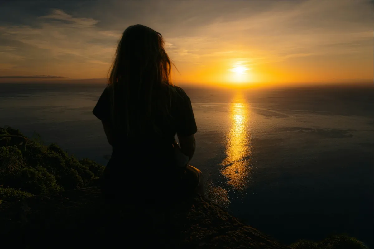 Silhouette of a woman meditating on a cliffside during a golden sunset, symbolizing the intentional practice of raising emotional frequency and nervous system regulation