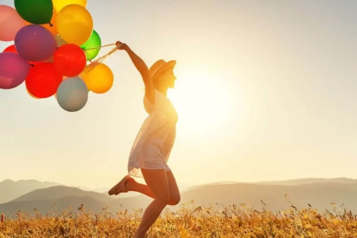 A woman running joyfully through a golden field with vibrant balloons, symbolizing the milestone celebration and tangible change of a higher energy awakening.