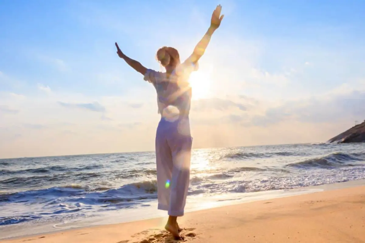 A woman in white standing on a beach at sunrise with arms raised, representing the clear signs your vibration is rising and the flow of a higher energy awakening.